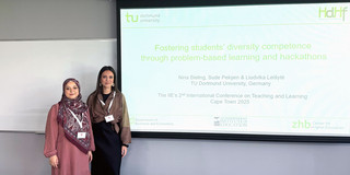 Foto: Sude Pekşen und Nina Bieling stehen in einem Konferenzraum, neben ihnen hängt eine Leinwand mit ihrer Präsentation.